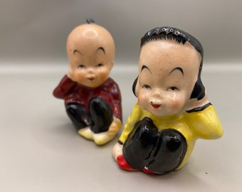 May include: Two ceramic figurines of children sitting with their legs crossed. The figurine on the left is wearing a red shirt and black pants. The figurine on the right is wearing a yellow shirt and black pants.