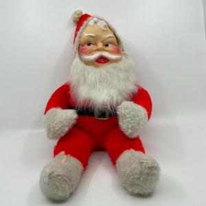 May include: A vintage Santa Claus doll featuring a molded plastic face with painted features, a long white faux fur beard, and a red plush suit. The doll wears a matching red hat with a white pom-pom, a black belt with a buckle, and white plush trim.