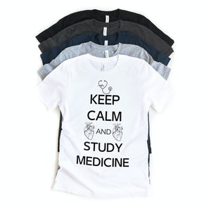 May include: White t-shirt with black text that reads "Keep Calm and Study Medicine". The text is accompanied by a stethoscope and two hearts. The shirt is stacked on top of other t-shirts in various colors.