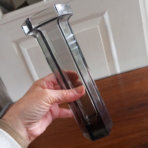 May include: A clear glass vase with a geometric design. The vase is tall and narrow, with a rectangular base and a square top.