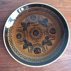 May include: A round, brown ceramic plate with a decorative floral and geometric pattern. The plate features a dark brown rim and a lighter brown center, with black floral and geometric designs. The plate is likely vintage or retro.