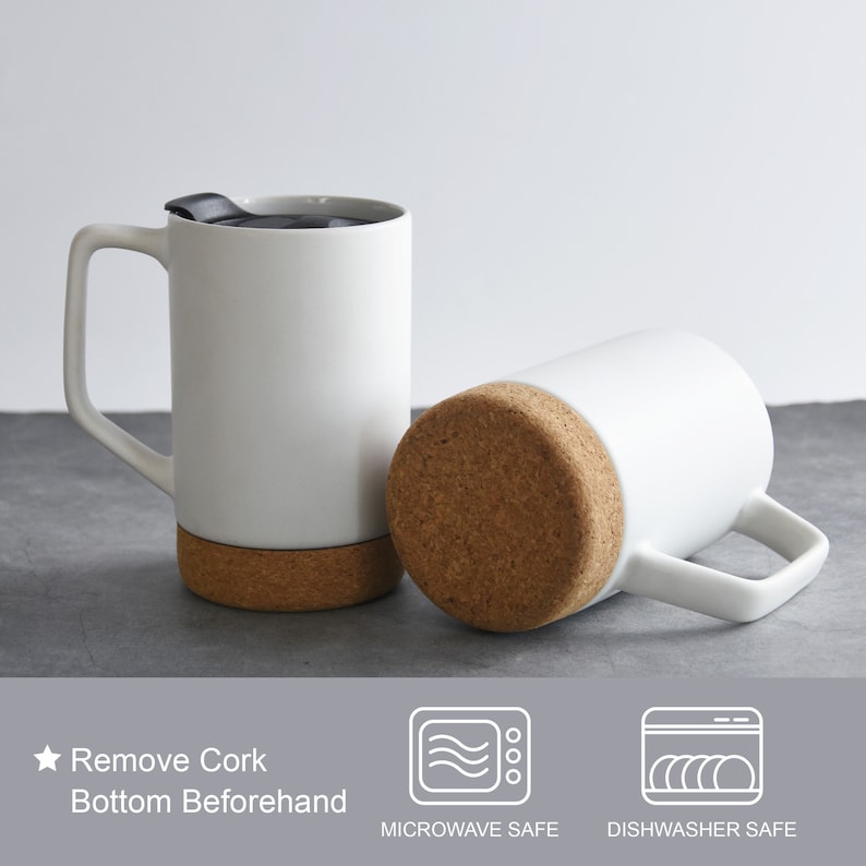 May include: Two white ceramic mugs with cork bottoms. The mugs have a black lid and a handle. The text "Remove Cork Bottom Beforehand" is visible below the mugs. The text "Microwave Safe" and "Dishwasher Safe" is visible below the mugs.