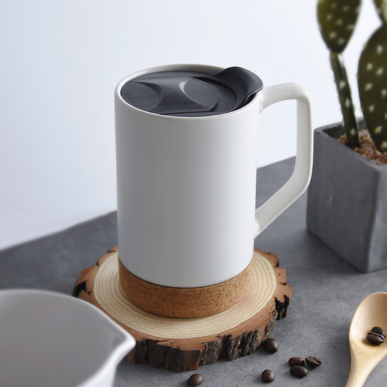 May include: White ceramic mug with a black lid and a cork base. The mug has a handle and is sitting on a wooden coaster.