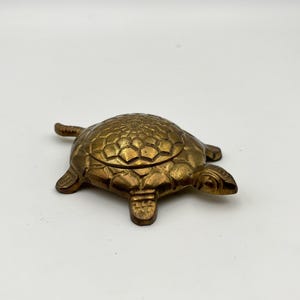 May include: A brass turtle figurine featuring a detailed, textured shell pattern on its back. The object has four legs, a small head, and a short tail, with a metallic gold-toned finish and visible seams indicating a hinged lid construction.