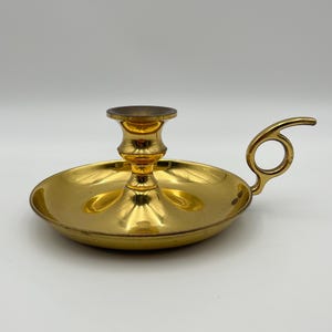 Puede incluir: Un candelabro de latón pulido en tono dorado con una base circular ancha que sirve como bandeja para gotas y un soporte central para una vela. La pieza incluye un asa curva con un orificio circular para el dedo, facilitando su transporte.