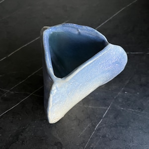 Könnte beinhalten: Eine handgefertigte Keramikvase mit einer einzigartigen, asymmetrischen Form. Die Vase zeigt einen Farbverlauf von Blautönen, der von einem helleren Farbton am Boden zu einem tieferen Blau im Inneren übergeht. Die strukturierte Oberfläche trägt zu ihrer künstlerischen Ausstrahlung bei.