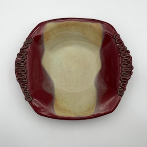 Op de afbeelding: Een vierkante keramische kom met afgeronde hoeken, voorzien van een glanzend dieprood glazuur aan de buitenkant en randen, en een gladde, lichtbeige binnenkant. Twee decoratieve, verhoogde, golvende kleislierten zijn aan de zijkanten bevestigd.