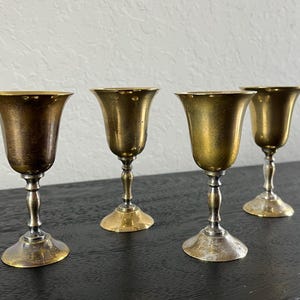 Puede incluir: Cuatro vasos de chupito de metal de color dorado con base plateada. Los vasos están dispuestos en fila sobre una superficie negra.