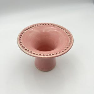 May include: A light pink ceramic flower frog or vase featuring a wide, flared rim with a series of evenly spaced circular holes around the edge. The object has a smooth, glossy finish and a cylindrical base that tapers slightly toward the center.