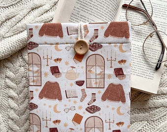 May include: A white book sleeve with a brown and white pattern of stars, moons, books, candles, teacups, butterflies, and sweaters. The sleeve has a brown fabric tag with the text "tula makes".