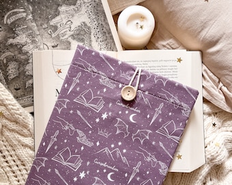 May include: A purple fabric book sleeve featuring a white line-art pattern of daggers, open books, bat wings, crowns, stars, and the word Cursebreaker. The sleeve has a wooden button closure with a white cord and a small rectangular brand label.