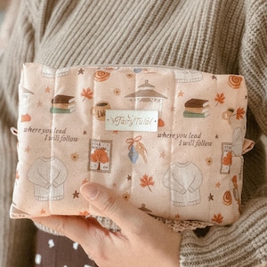 Puede incluir: Un estuche acolchado rectangular de color melocotón claro con un patrón repetitivo de ilustraciones otoñales, que incluyen jerséis, libros, tazas de café y cenadores. El texto "where you lead I will follow" y "Fairy Tula" está impreso en la parte frontal.