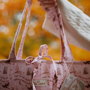 May include: A light pink quilted tote bag featuring a repeating pattern of dark brown dragons, castles, crescent moons, and stars. The fabric includes the text "I had the time of my life fighting dragons with you" and a "Tula Makes" brand label.