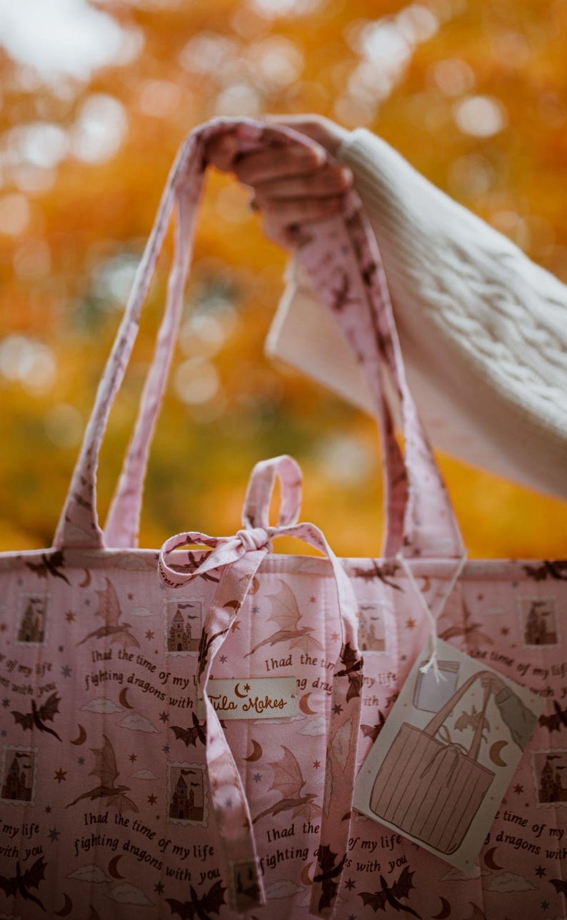 May include: A light pink quilted tote bag featuring a repeating pattern of dark brown dragons, castles, crescent moons, and stars. The fabric includes the text "I had the time of my life fighting dragons with you" and a "Tula Makes" brand label.