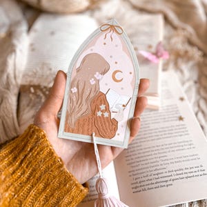 Może przedstawiać: Zakładka do książki z ilustracją osoby czytającej, z półksiężycem i gwiazdami. Ma różowy chwost i jest trzymana nad otwartą książką. Ilustracja wykorzystuje miękkie, stonowane kolory.
