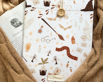 Puede incluir: Una funda de tela blanca para libros con un patrón repetitivo de iconos mágicos en marrón y dorado, incluyendo gafas, una varita, una bufanda, un búho y cartas. La funda se cierra con un botón de madera y tiene una etiqueta que dice tula makes.