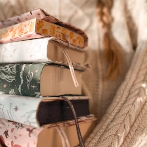 Może przedstawiać: Stos książek w twardej oprawie z dekoracyjnymi okładkami. Książki mają różne wzory, w tym wzory kwiatowe i motywy niebieskie. Każda książka ma wstążkową zakładkę. Książki są trzymane przed kremowym swetrem z warkoczowym splotem.