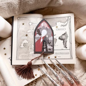 Pode incluir: Um marcador de página com uma mulher em um manto vermelho em pé em um arco gótico com uma lua crescente e estrelas no fundo. O marcador de página é feito de papel e tem uma borla.