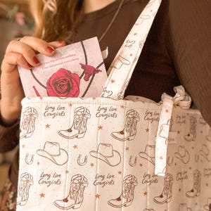 May include: A quilted tote bag in off-white featuring a repeating brown print of cowboy boots, hats, horseshoes, and stars. The text "Long Live Cowgirls" is printed throughout the pattern. A card with a red rose graphic is tucked inside.