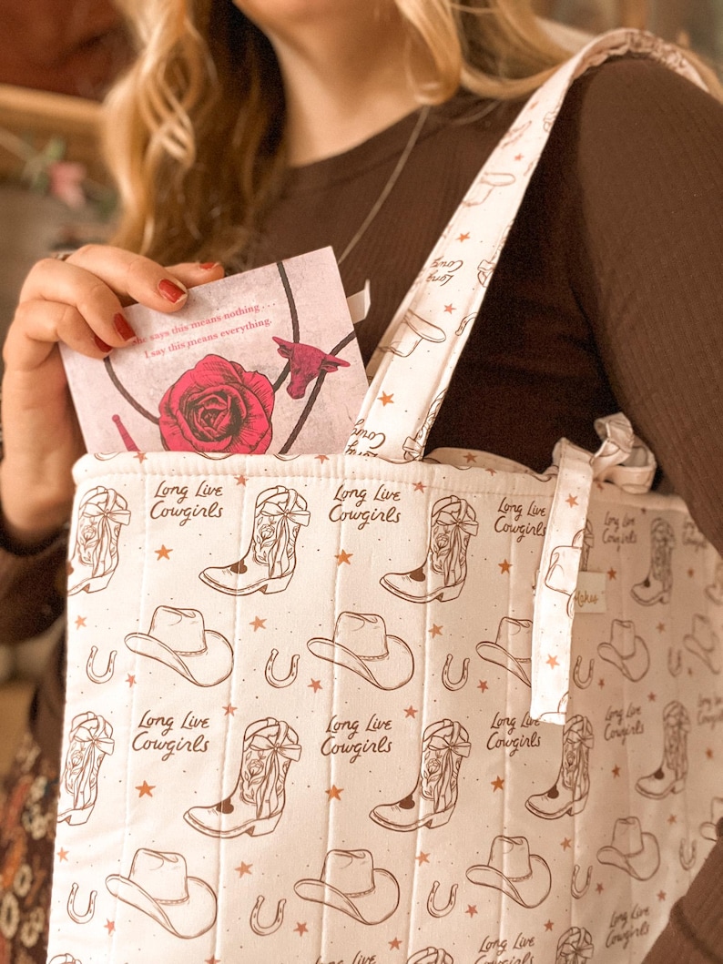 May include: A quilted tote bag in off-white featuring a repeating brown print of cowboy boots, hats, horseshoes, and stars. The text "Long Live Cowgirls" is printed throughout the pattern. A card with a red rose graphic is tucked inside.