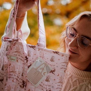 May include: A quilted tote bag in light pink featuring a repeating pattern of brown dragons, crescent moons, stars, and castles. The fabric includes the text "I had the time of my life fighting dragons with you" and a small tied bow on the handle.