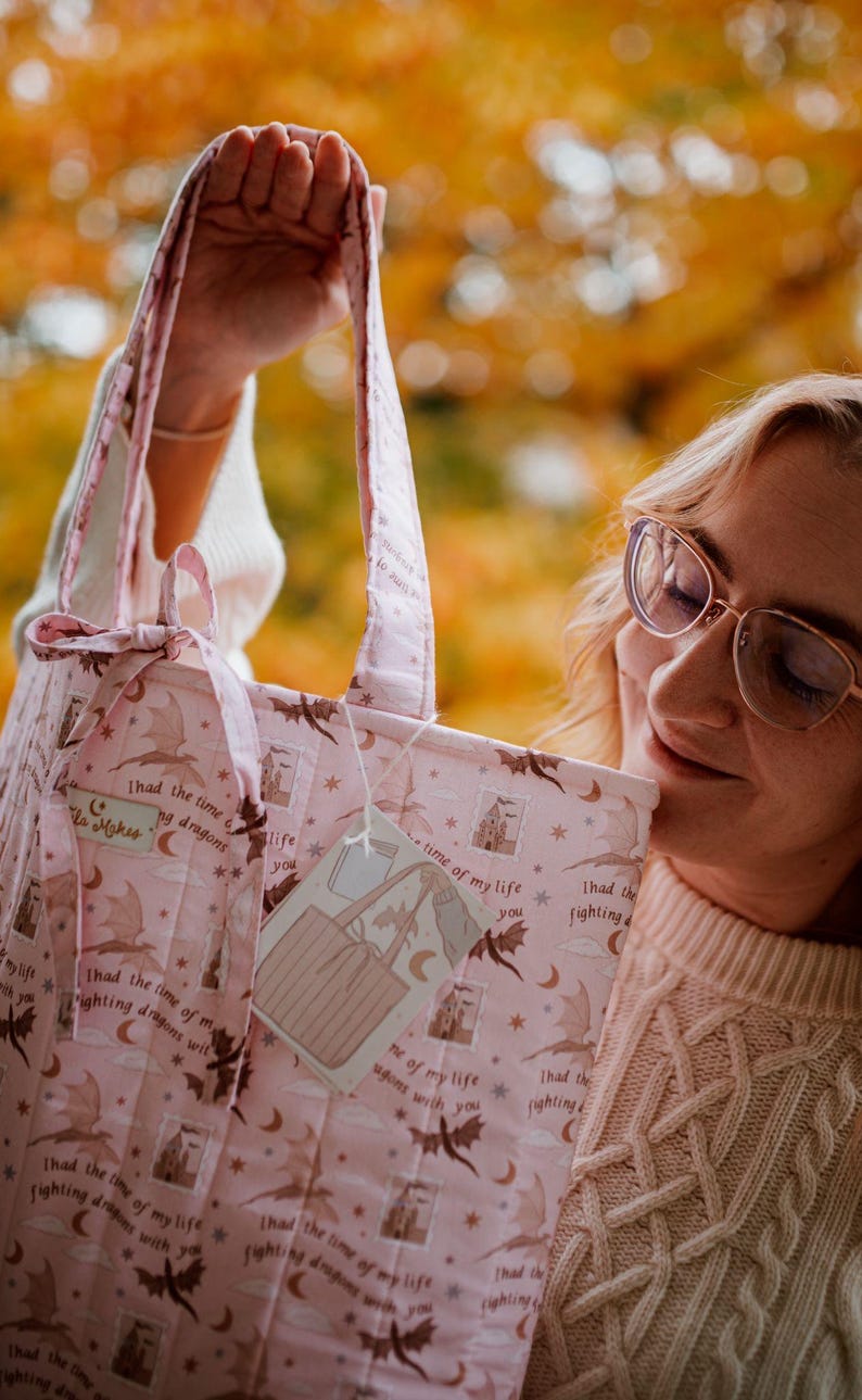 May include: A quilted tote bag in light pink featuring a repeating pattern of brown dragons, crescent moons, stars, and castles. The fabric includes the text "I had the time of my life fighting dragons with you" and a small tied bow on the handle.