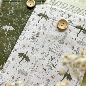 Puede incluir: Dos fundas de tela para libros, una en verde oliva y otra en blanco, con un patrón de espadas, coronas, dragones, pájaros y estrellas. Cada funda tiene un botón de madera con el texto "Handmade with love" y una etiqueta.