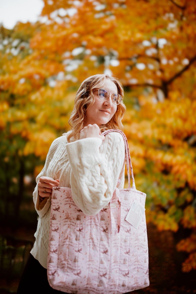 May include: A light pink quilted tote bag featuring a repeating pattern of small dark silhouettes of flying dragons, crescent moons, and stars, with two matching fabric shoulder straps and a small white rectangular tag attached to the side.