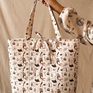 May include: A quilted tote bag in a light cream color featuring a repeating dark brown pattern of mermaids, typewriters, ghosts, keys, envelopes, and butterflies. The bag includes two shoulder straps and a small fabric label reading Tula Makes.