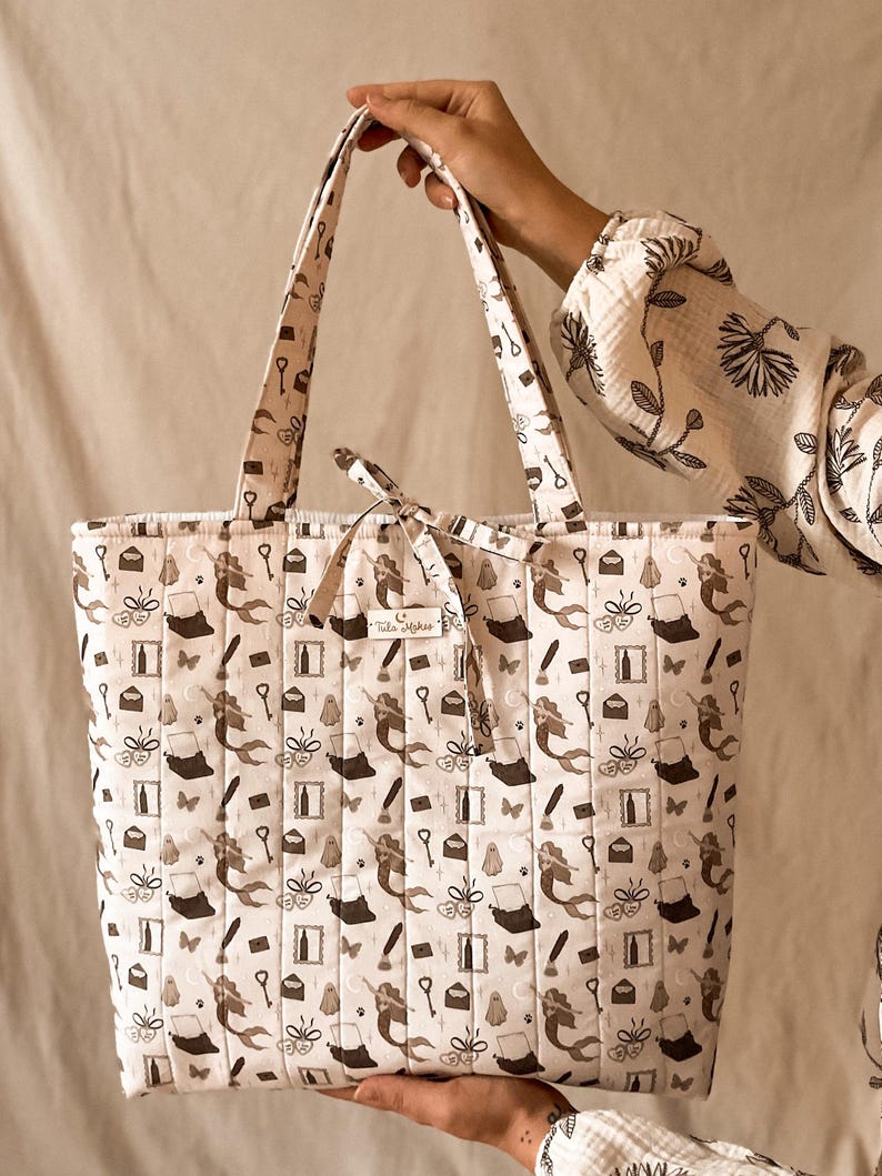May include: A quilted tote bag in a light cream color featuring a repeating dark brown pattern of mermaids, typewriters, ghosts, keys, envelopes, and butterflies. The bag includes two shoulder straps and a small fabric label reading Tula Makes.