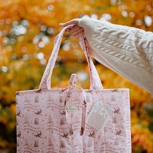 May include: A light pink quilted tote bag featuring a repeating pattern of dark brown flying dragons, crescent moons, stars, and the text "Had the time of my life fighting dragons with you." The bag has two top handles and a small tie closure at the top.