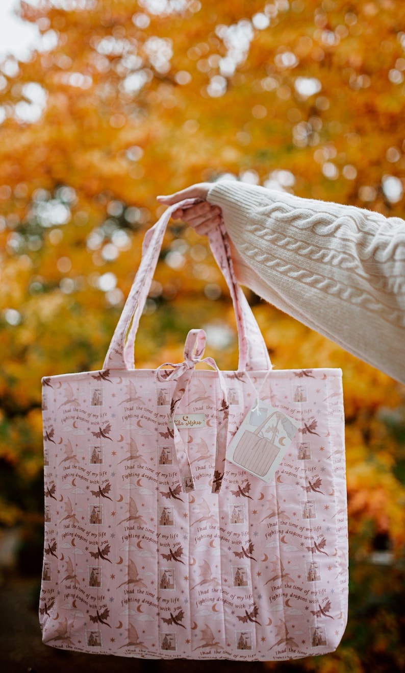 May include: A light pink quilted tote bag featuring a repeating pattern of dark brown flying dragons, crescent moons, stars, and the text "Had the time of my life fighting dragons with you." The bag has two top handles and a small tie closure at the top.