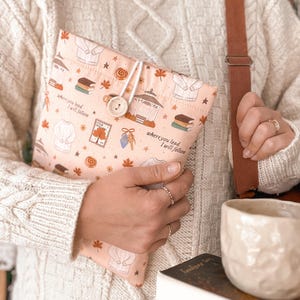 Pode incluir: Uma bolsa de tecido em tom pêssego claro com um padrão repetitivo de camisolas, coretos, livros e folhas de outono. O texto "where you lead I will follow" está impresso na frente. A bolsa é fechada com um cordão branco e um botão de madeira.