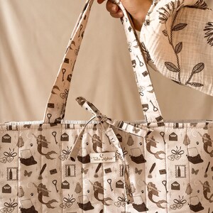 May include: A quilted tote bag in a light beige fabric featuring a repeating dark brown pattern of mermaids, typewriters, keys, ghosts, envelopes, and butterflies. A small rectangular label on the front displays the text Tula Makes.