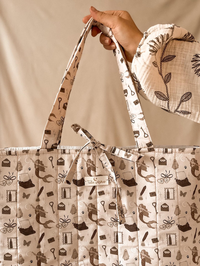 May include: A quilted tote bag in a light beige fabric featuring a repeating dark brown pattern of mermaids, typewriters, keys, ghosts, envelopes, and butterflies. A small rectangular label on the front displays the text Tula Makes.