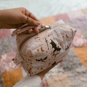 Pode incluir: Uma bolsa acolchoada rosa-claro com fecho de correr, apresentando um padrão repetitivo de silhuetas pretas de dragões, espadas, luas crescentes, estrelas e nuvens brancas. O texto "DRAGON RIDER" está impresso a preto no tecido perto do fecho.