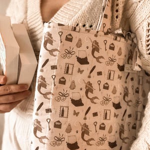 May include: A quilted tote bag in a light beige fabric featuring a repeating dark brown pattern of mermaids, typewriters, ghosts, keys, envelopes, and hearts. The bag has two fabric handles and a small rectangular tag on the side seam.