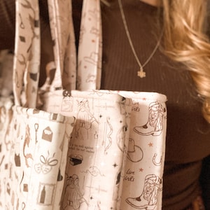 May include: A cream-colored fabric tote bag featuring a repeating brown line-art pattern of cowboy boots, hats, horseshoes, stars, and figures holding swords. The text "Live girls" is printed in a script font alongside various small icons.