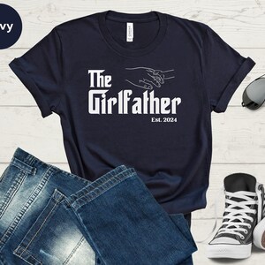 May include: Navy blue t-shirt with white text that reads "The Girlfather Est. 2024". The text is in a bold, stylised font. The shirt features a graphic of two hands holding each other.