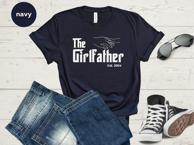 May include: Navy blue t-shirt with white text that reads "The Girlfather Est. 2024". The text is in a bold, stylised font. The shirt features a graphic of two hands holding each other.