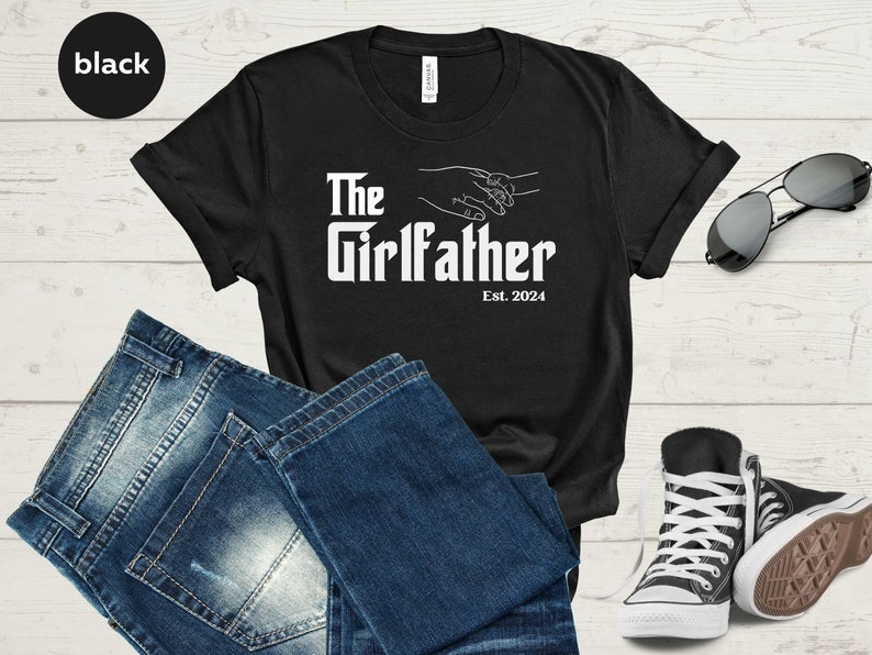May include: Black t-shirt with white text that reads "The Girlfather Est. 2024" and a graphic of two hands holding each other.