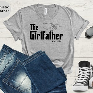 May include: Grey t-shirt with the text "The Girlfather Est. 2024" and a graphic of two hands holding each other.