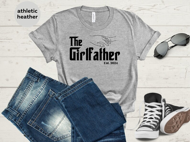 May include: Grey t-shirt with the text "The Girlfather Est. 2024" and a graphic of two hands holding each other.