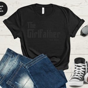 May include: Black t-shirt with the text "The Girlfather Est. 2023" printed on it. The shirt is laid out on a wooden surface with a pair of jeans, sunglasses, and trainers.