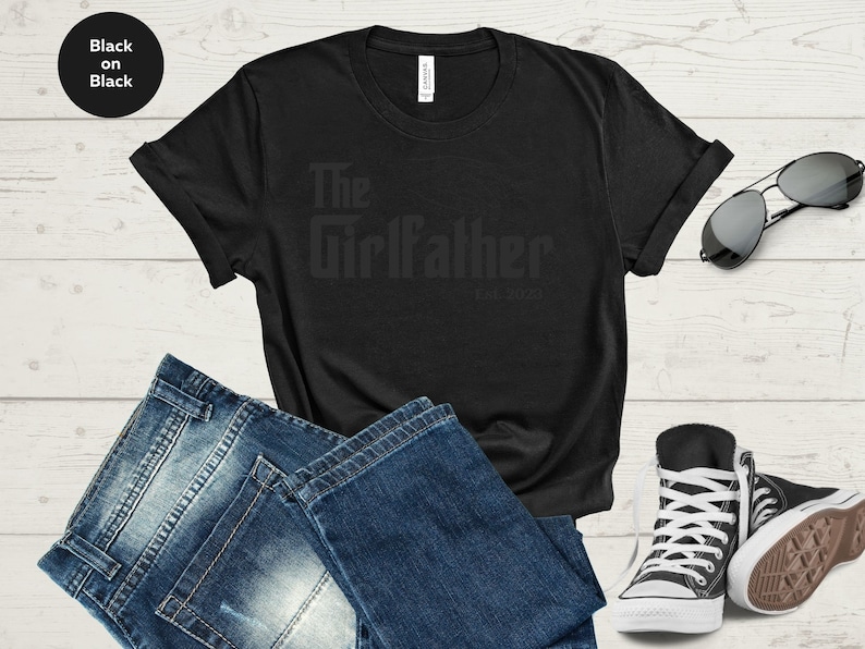 May include: Black t-shirt with the text "The Girlfather Est. 2023" printed on it. The shirt is laid out on a wooden surface with a pair of jeans, sunglasses, and trainers.
