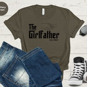 May include: A military green t-shirt with the text "The Girlfather Est. 2024" in black. The shirt features a graphic of two hands holding each other.