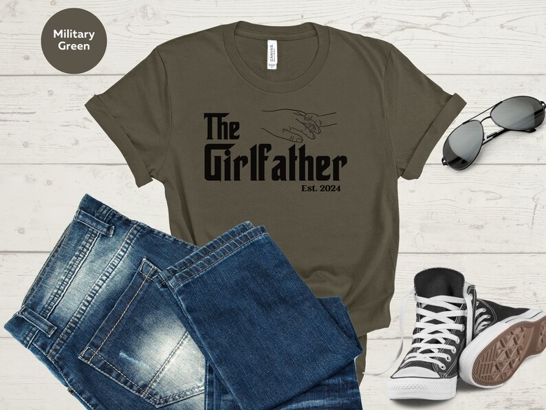 May include: A military green t-shirt with the text "The Girlfather Est. 2024" in black. The shirt features a graphic of two hands holding each other.