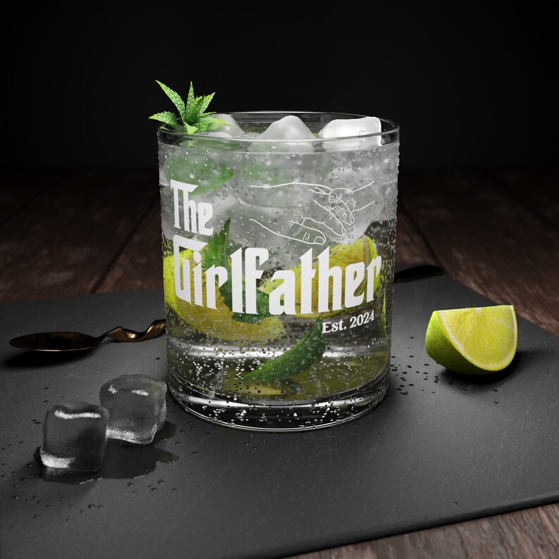 Peut inclure: Un verre transparent avec une inscription blanche qui dit "The Girlfather Est. 2024". Le verre est rempli de glace, d'un quartier de citron vert et d'une brindille de menthe.
