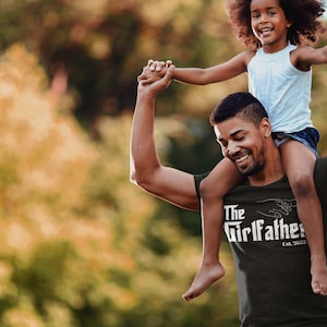 May include: A man is carrying a young girl on his shoulders. He is wearing a black t-shirt with the text "The Girlfather Est. 2022" in white. The girl is wearing a light blue tank top and denim shorts. They are both smiling and looking at the camera.
