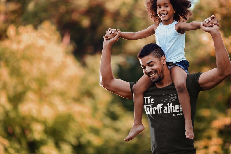 May include: A man is carrying a young girl on his shoulders. He is wearing a black t-shirt with the text "The Girlfather Est. 2022" in white. The girl is wearing a light blue tank top and denim shorts. They are both smiling and looking at the camera.